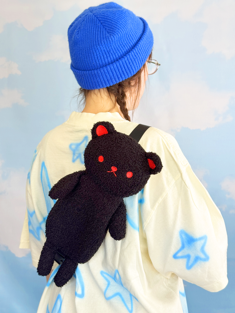 Person wearing a blue beanie and holding a black cat-shaped backpack against a light blue starry background