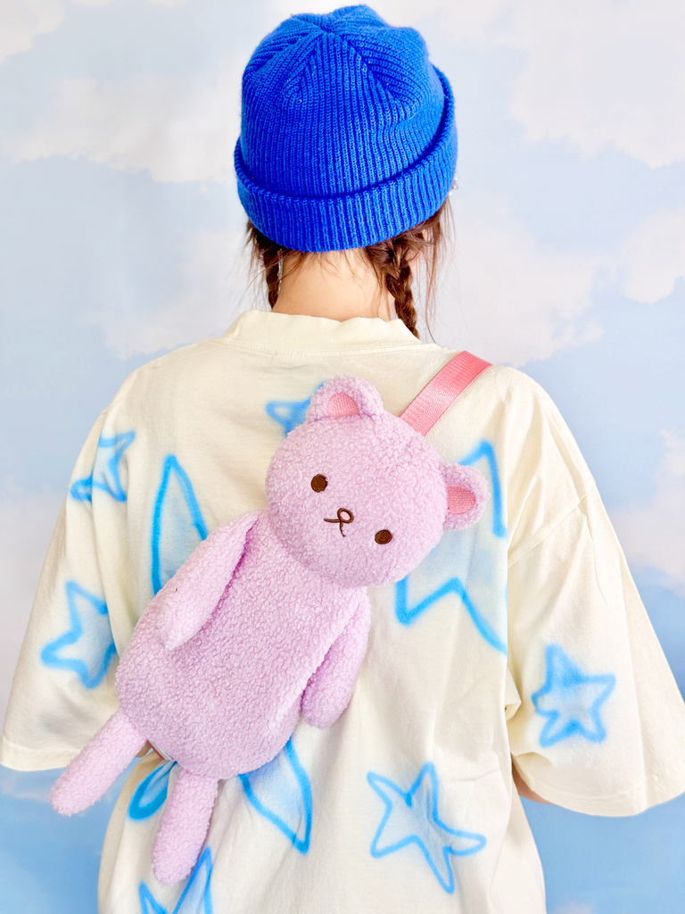 Person wearing a blue beanie and a white jacket with star patterns, holding a pink plush toy against a light blue background.